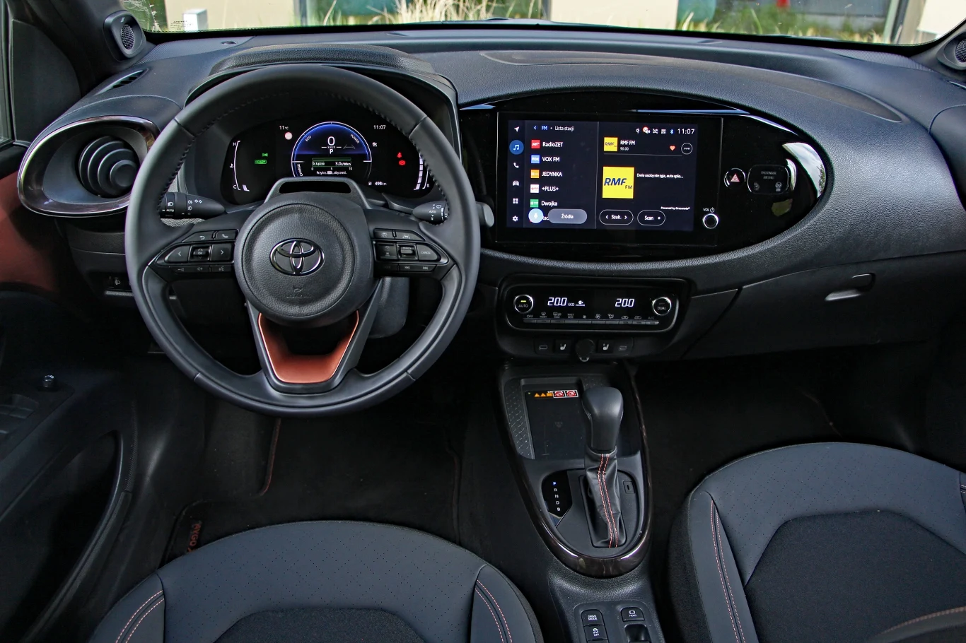 Toyota Aygo X Hybrid ma zaskakująco bogate wyposażenie jak na segment A, ale o przynależności do niego przypominają liczne oszczędności - takie jak brak centralnych nawiewów Wnętrze samochodu marki Toyota z nowoczesną kierownicą wielofunkcyjną, cyfrowym wyświetlaczem za kierownicą, dużym ekranem dotykowym systemu multimedialnego na środkowej konsoli i automatyczną skrzynią biegów.