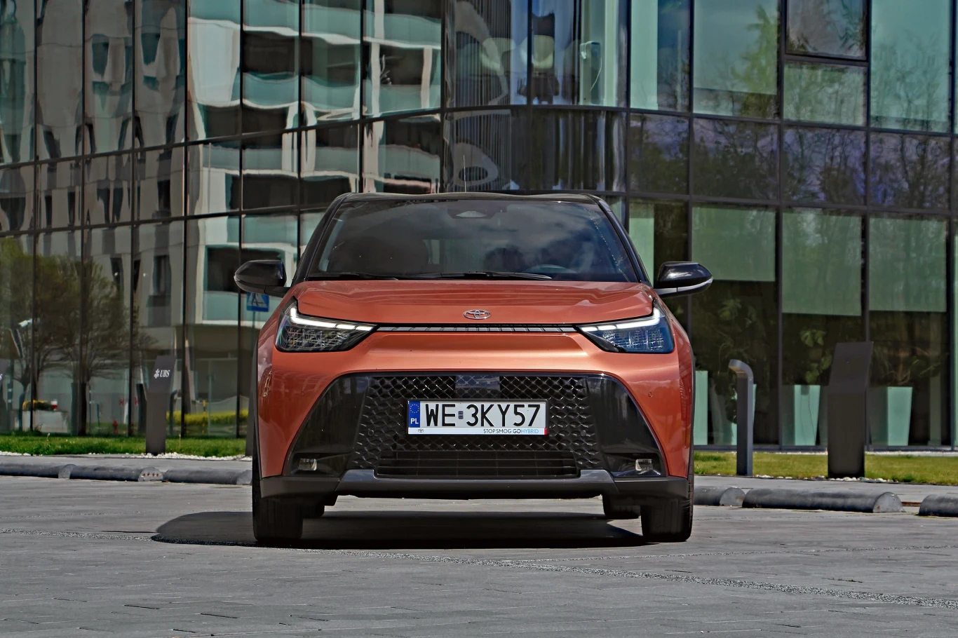 Toyota Aygo X Hybrid Czerwony miejski samochód elektryczny ustawiony na tle nowoczesnego przeszklonego budynku.
