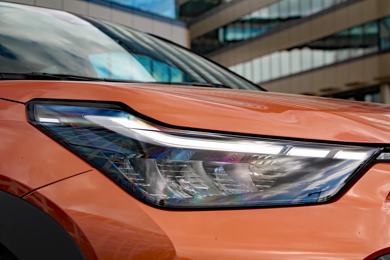 Toyota Aygo X Hybrid Przedni reflektor nowoczesnego, pomarańczowego samochodu z wyraźnie zarysowaną linią nadwozia i fragmentem szyby w tle.