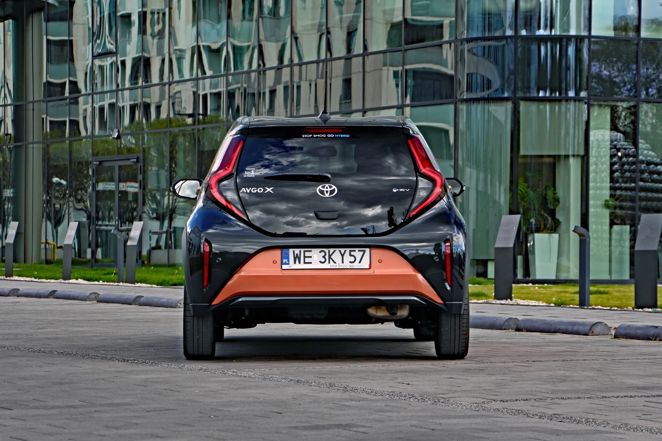 Toyota Aygo X Hybrid Czarny miejski samochód Toyota Aygo X z pomarańczowymi akcentami stoi zaparkowany tyłem przy nowoczesnym, szklanym budynku.