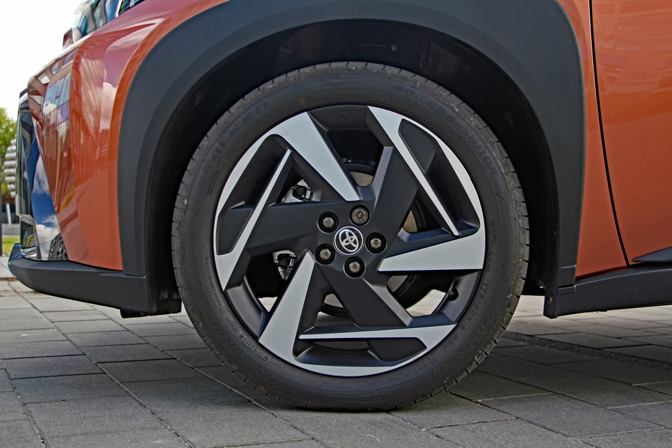 Toyota Aygo X Hybrid Nowoczesna, dwukolorowa felga aluminiowa z logo marki Toyota na oponie zamontowanej w przednim nadkolu pomarańczowego samochodu osobowego.