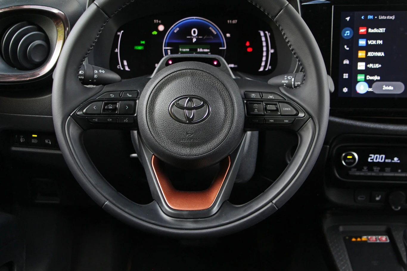 Toyota Aygo X Hybrid Kierownica Toyoty z przyciskami sterującymi, cyfrowy wyświetlacz deski rozdzielczej pokazujący prędkościomierz i wskaźniki oraz fragment nowoczesnego systemu multimedialnego z wyświetlaczem dotykowym po prawej stronie.