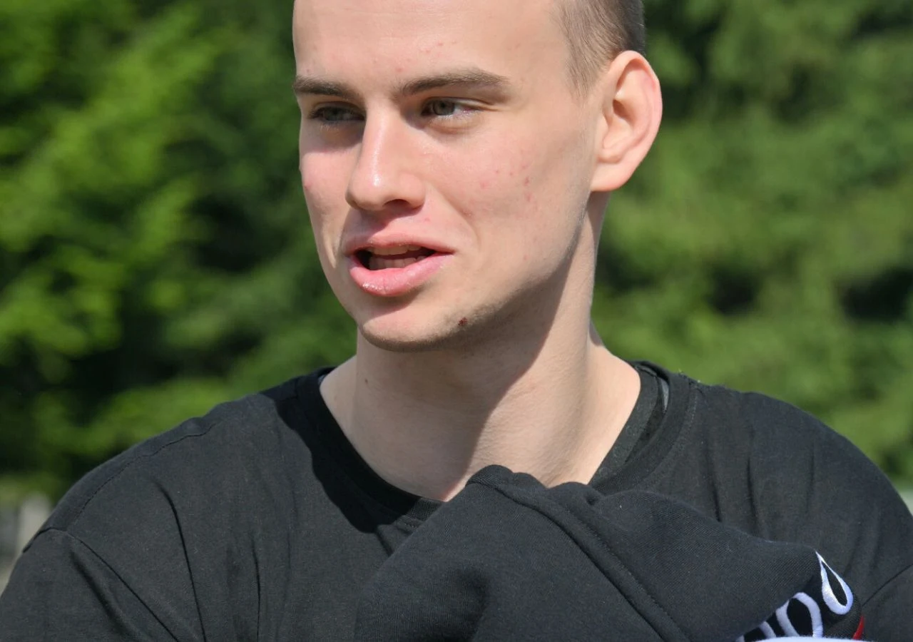 Łatwogang Influencer Łatwogang, czyli Piotr Hancke, w czarnej koszulce, stoi na tle zieleni.