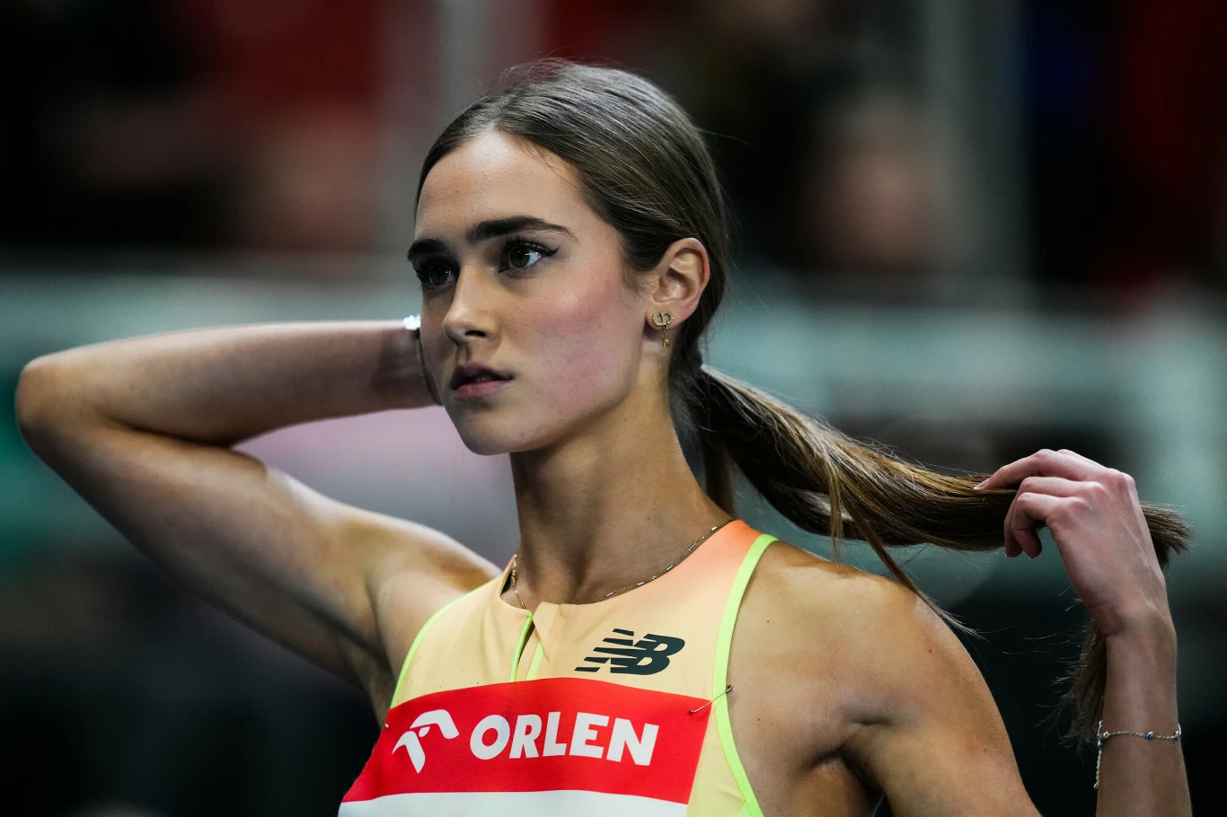 Lekkoatletka w sportowej koszulce z numerem startowym i logotypem ORLEN, odgarniająca włosy dłonią podczas przygotowań do biegu.