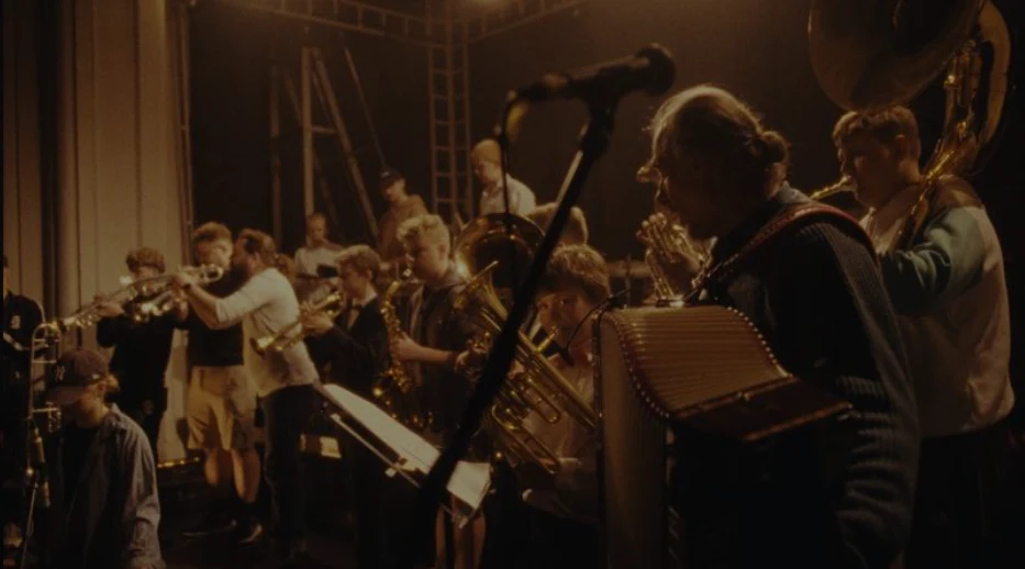 Grupa muzyków grająca na scenie, wśród instrumentów widoczne trąbki, puzony, akordeon i tuba, wszyscy skoncentrowani na wspólnym występie.