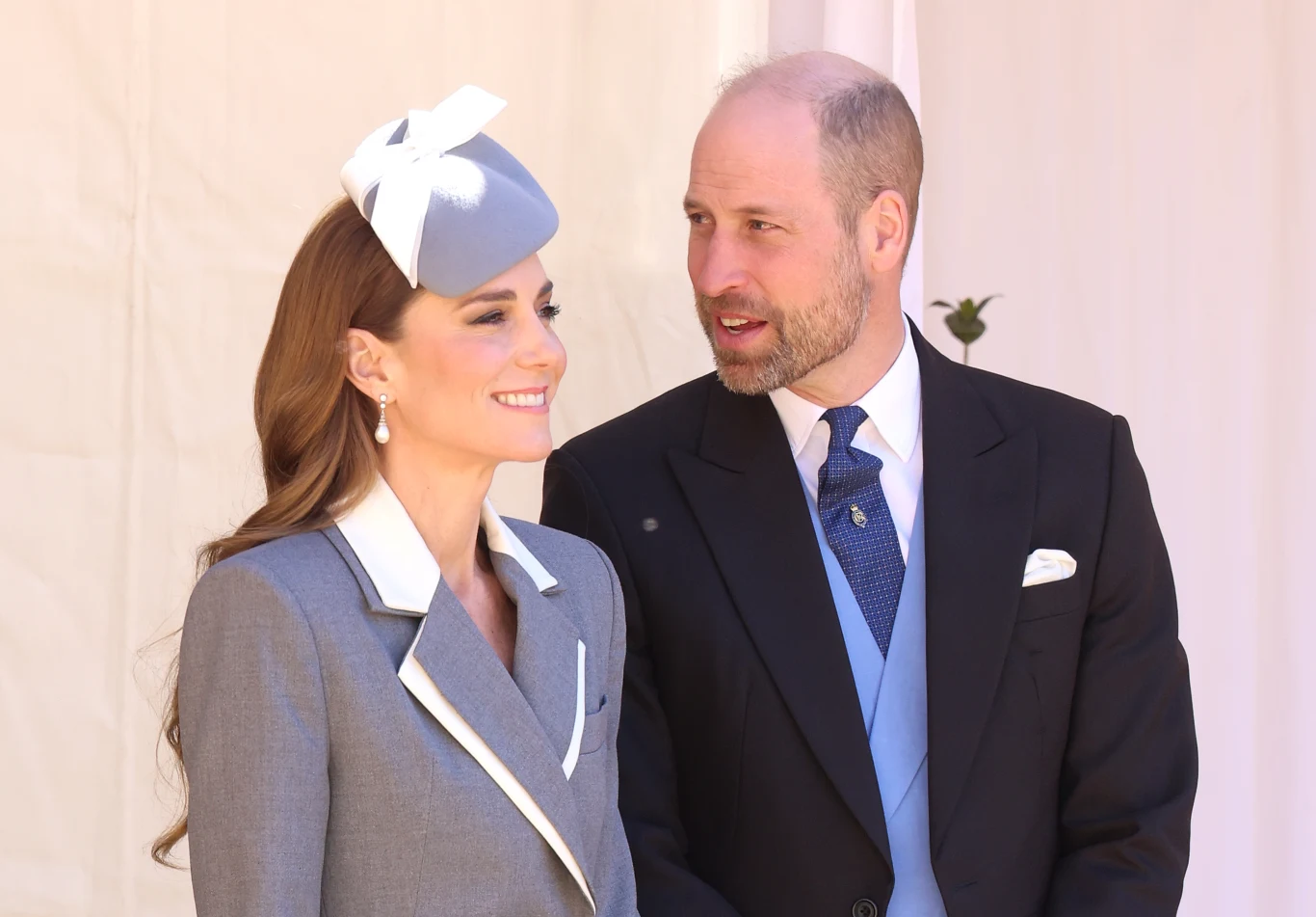 Elegancko ubrana kobieta w szarym kapeluszu z białą kokardą i kolczykami stoi obok mężczyzny w garniturze z jasnoniebieską kamizelką i niebieskim krawatem; oboje uśmiechnięci, zwróceni ku sobie.