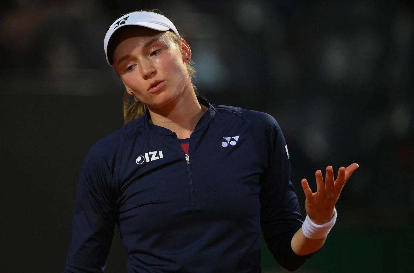 Jelena Rybakina Tenisistka ubrana w granatowy strój sportowy i białą czapkę z daszkiem wykonuje gest dłonią, mając wyraz twarzy sugerujący dezaprobatę lub zniechęcenie.