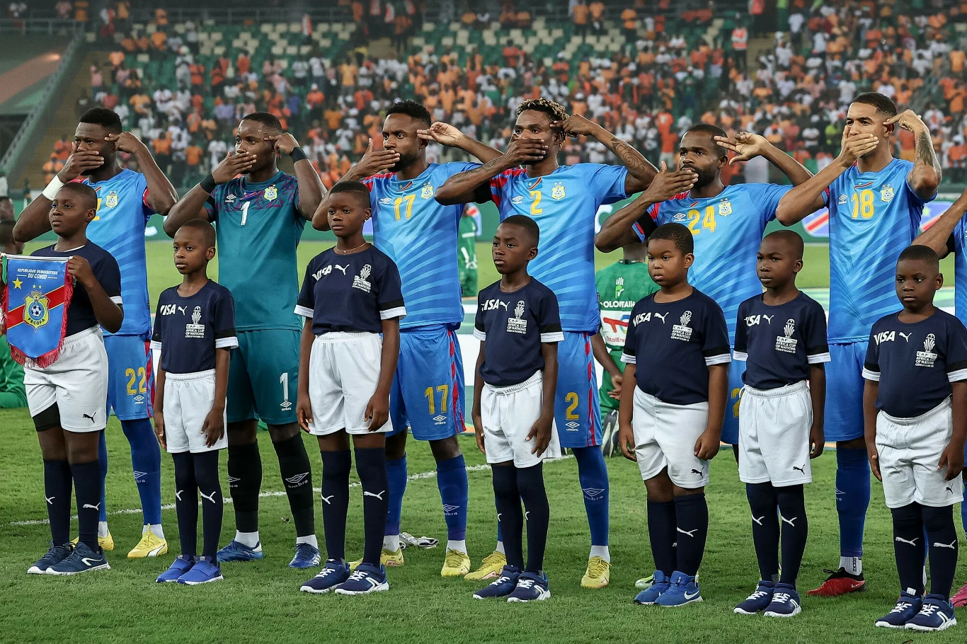 Piłkarze drużyny w niebieskich i żółtych strojach oraz dziecięca asysta stoją ramię w ramię, zawodnicy wykonują gest zakrywania ust, uszu i oczu na tle stadionu pełnego kibiców.