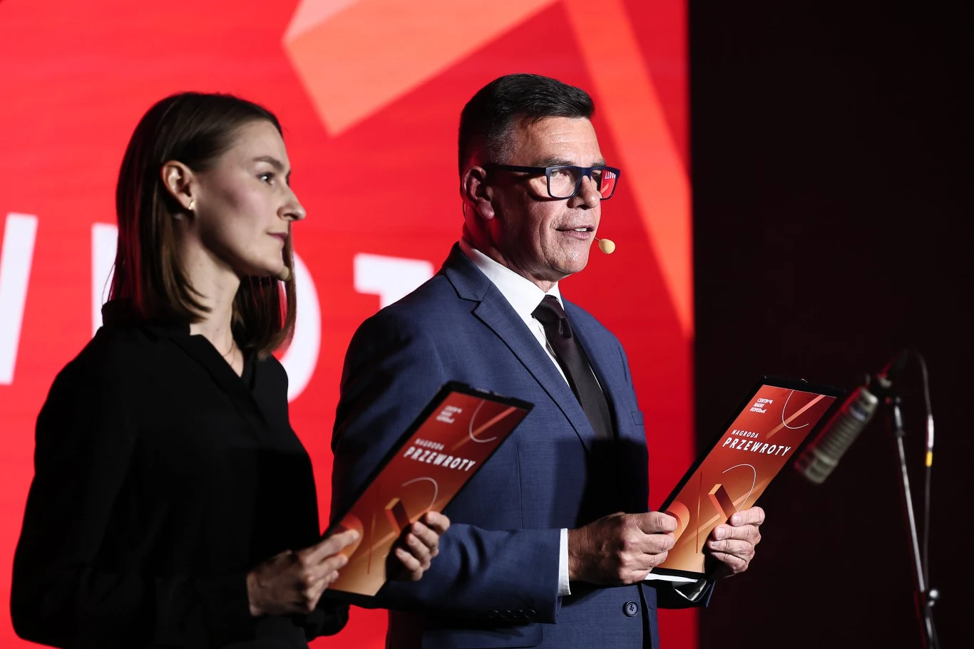 Dwoje elegancko ubranych prowadzących trzymających w rękach czerwone teczki z napisem podczas oficjalnego wydarzenia na tle jasnoczerwonego ekranu.