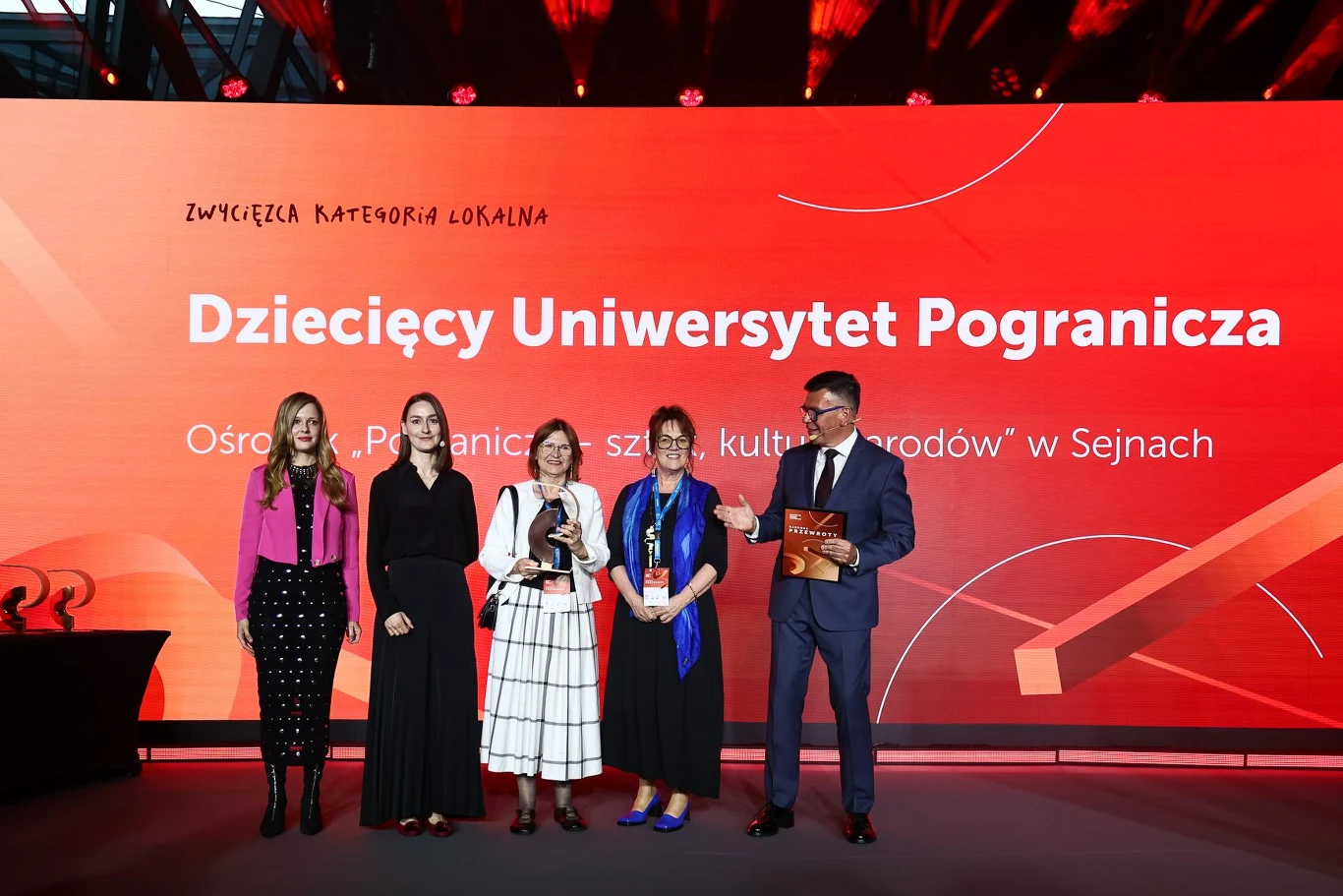 Pięć osób elegancko ubranych stoi na scenie z nagrodami na tle dużego ekranu z napisem „Dziecięcy Uniwersytet Pogranicza”, uśmiechając się do publiczności.