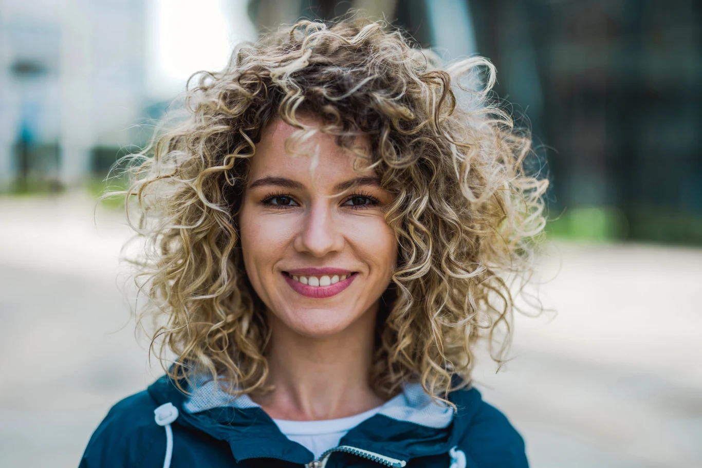 Uśmiechnięta kobieta z kręconymi blond włosami ubrana w granatową kurtkę, patrząca prosto w obiektyw.