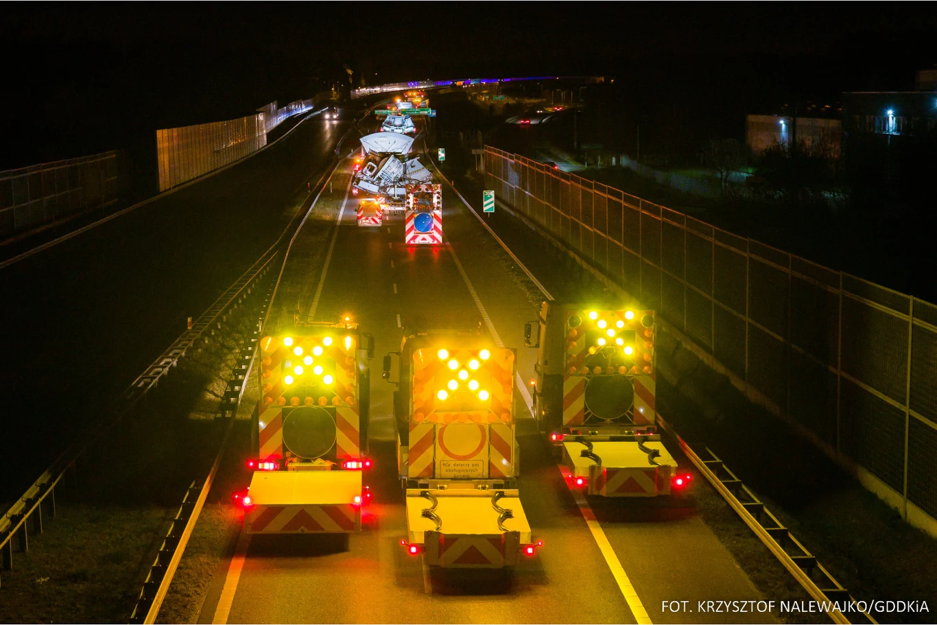 Kolumna samochodów wyposażonych w sygnalizację świetlną zabezpiecza prace drogowe na autostradzie nocą, ruch w stronę robót zablokowany, pozostałe pasy oddzielone barierami dźwiękochłonnymi.