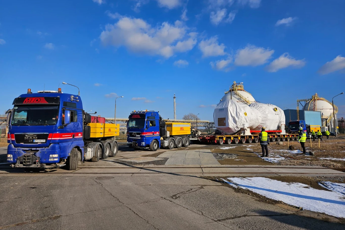 Konwój ciężarówek przewożących duży, osłonięty białą plandeką ładunek przemysłowy, eskortowany przez kilku pracowników w kamizelkach odblaskowych na terenie przemysłowym pod niebieskim niebem.