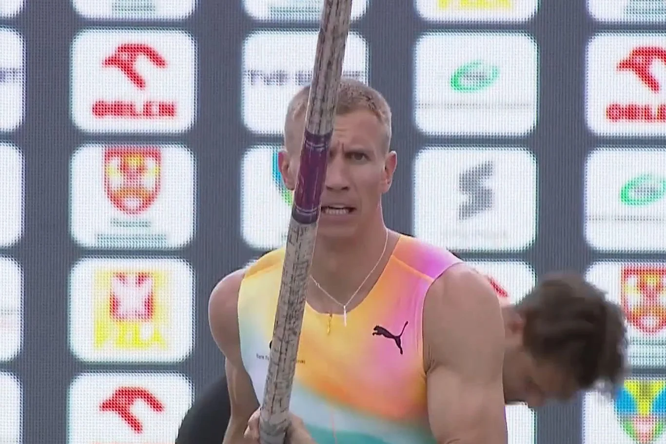 Zawodnik w kolorowej sportowej koszulce trzyma tyczkę do skoku o tyczce, w tle tablica z logotypami sponsorów.