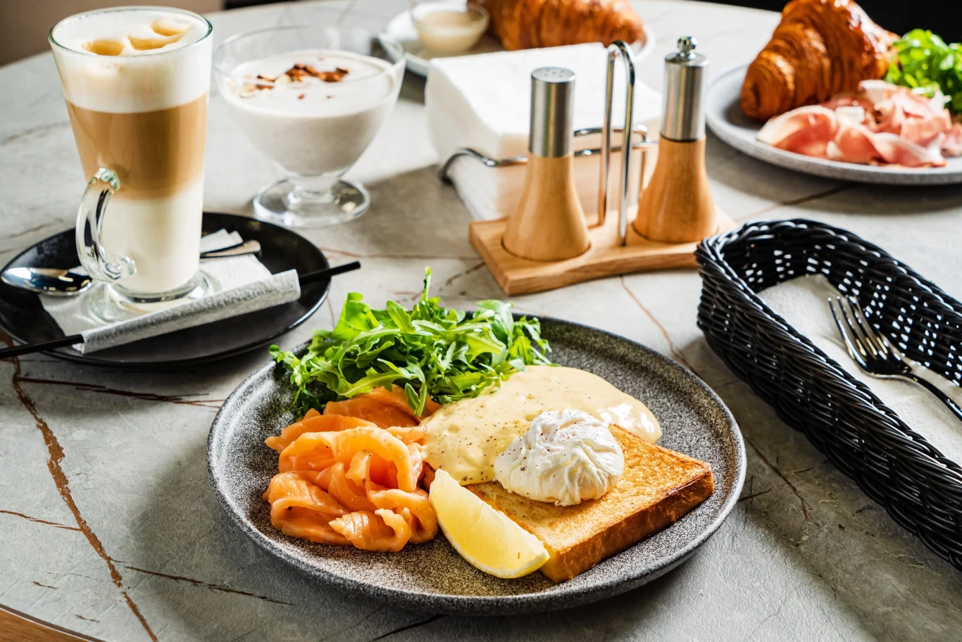 Tost z jajkiem w koszulce, sosem holenderskim, plastrami wędzonego łososia, rukolą i kawałkiem cytryny, obok kawa latte oraz jogurt w pucharku.