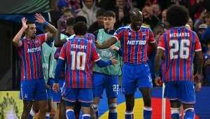 Piłkarze drużyny w pasiastych czerwono-niebieskich strojach celebrują zdobycie gola, otoczeni przez kolegów z zespołu na murawie stadionu.
