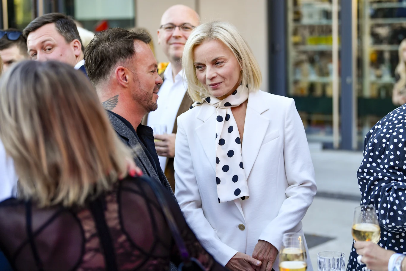 Kobieta w białym garniturze z apaszką w grochy rozmawia z mężczyzną o tatuażu na szyi podczas eleganckiego spotkania towarzyskiego na zewnątrz, wokół trzymane są kieliszki z winem.