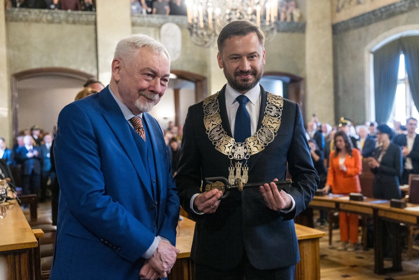 Dwaj byli prezydenci Krakowa: Jacek Majchrowski i Aleksander Miszalski Dwaj elegancko ubrani mężczyźni stoją obok siebie w sali urzędu podczas oficjalnej ceremonii, jeden z nich trzyma ozdobny łańcuch symbolizujący władzę miejską, w tle obecni są uczestnicy wydarzenia.