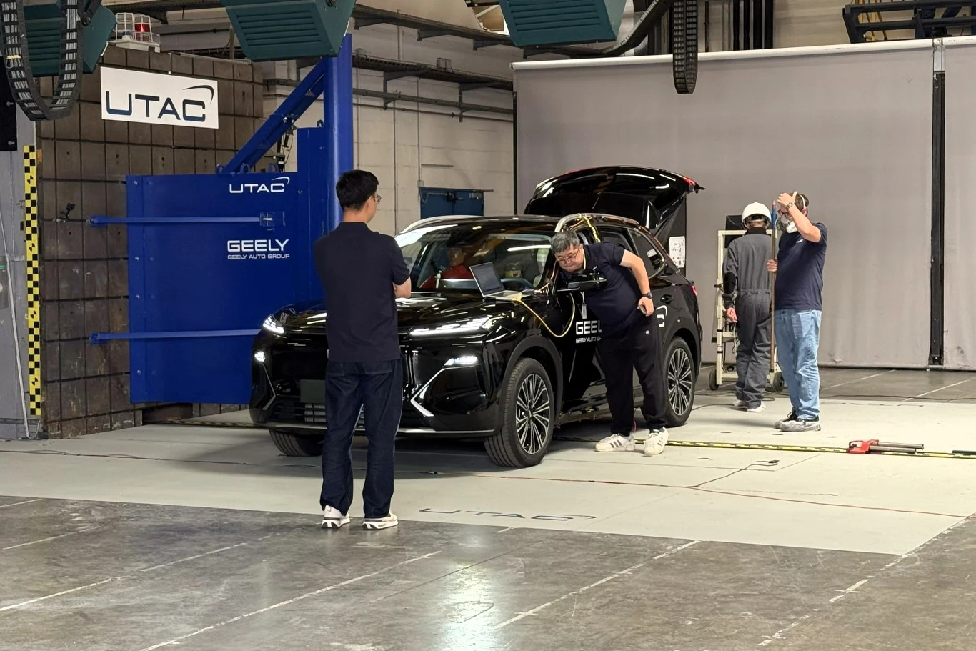 Czarny samochód marki Geely ustawiony w hali testowej, wokół którego pracują czterej technicy ubrani w kaski i ubrania robocze, przygotowujący pojazd do testów zderzeniowych.