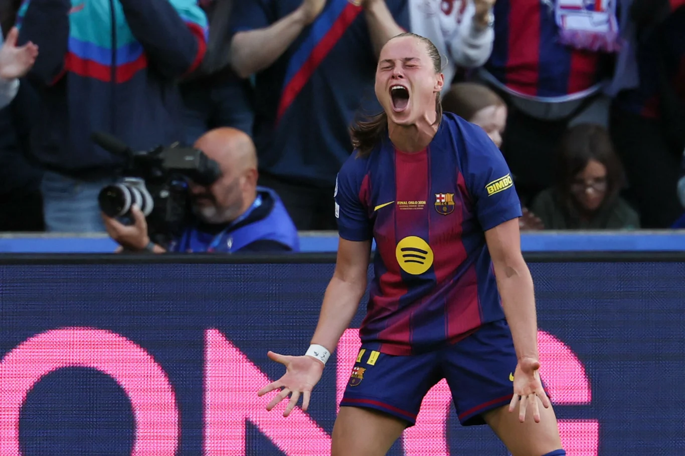 Piłkarka FC Barcelony w granatowo-bordowej koszulce wyraża silne emocje po zdobyciu gola, z szeroko otwartymi ustami i rozpostartymi rękami; w tle kibice dopingują drużynę.