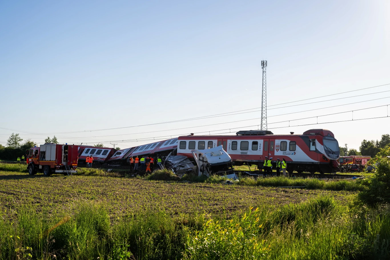 Wykolejony pociąg pasażerski z poważnie uszkodzonymi wagonami, strażacy i ratownicy pracujący przy wraku na środku pola, w tle wóz strażacki oraz infrastruktura kolejowa.