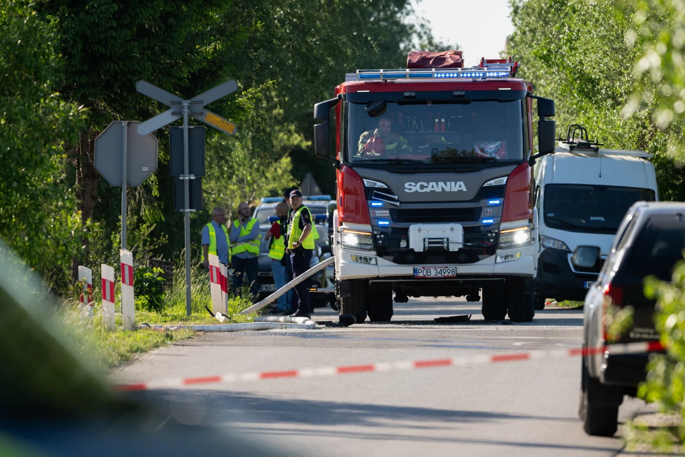 Wóz strażacki stojący na jezdni przy przejeździe kolejowym, obok grupy osób w kamizelkach odblaskowych prowadzących działania ratownicze; z prawej strony widoczny biały samochód dostawczy.