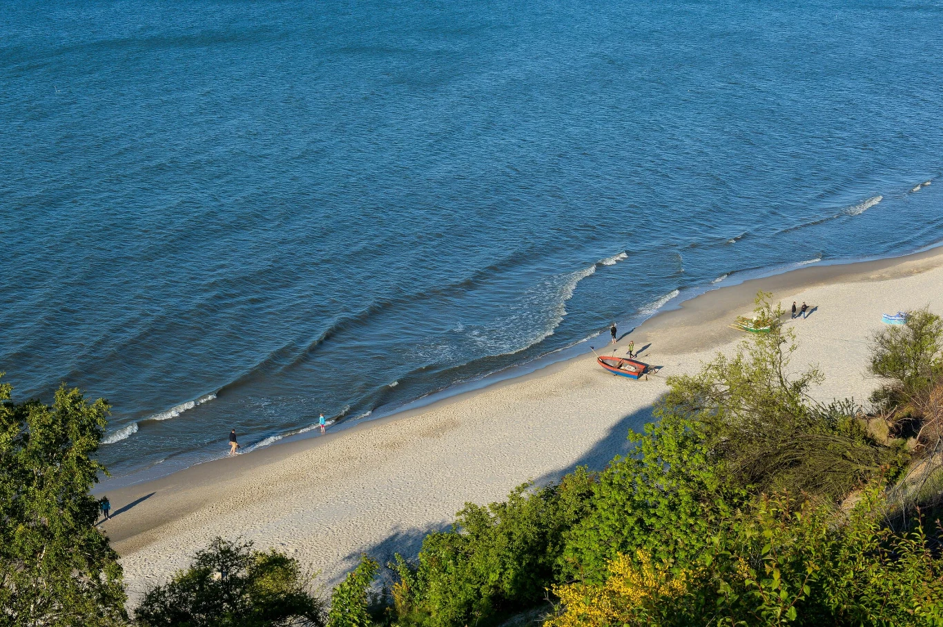Piaszczysta plaża nad brzegiem morza, kilka osób spaceruje wzdłuż linii wody, a przy brzegu stoi łódź rybacka.