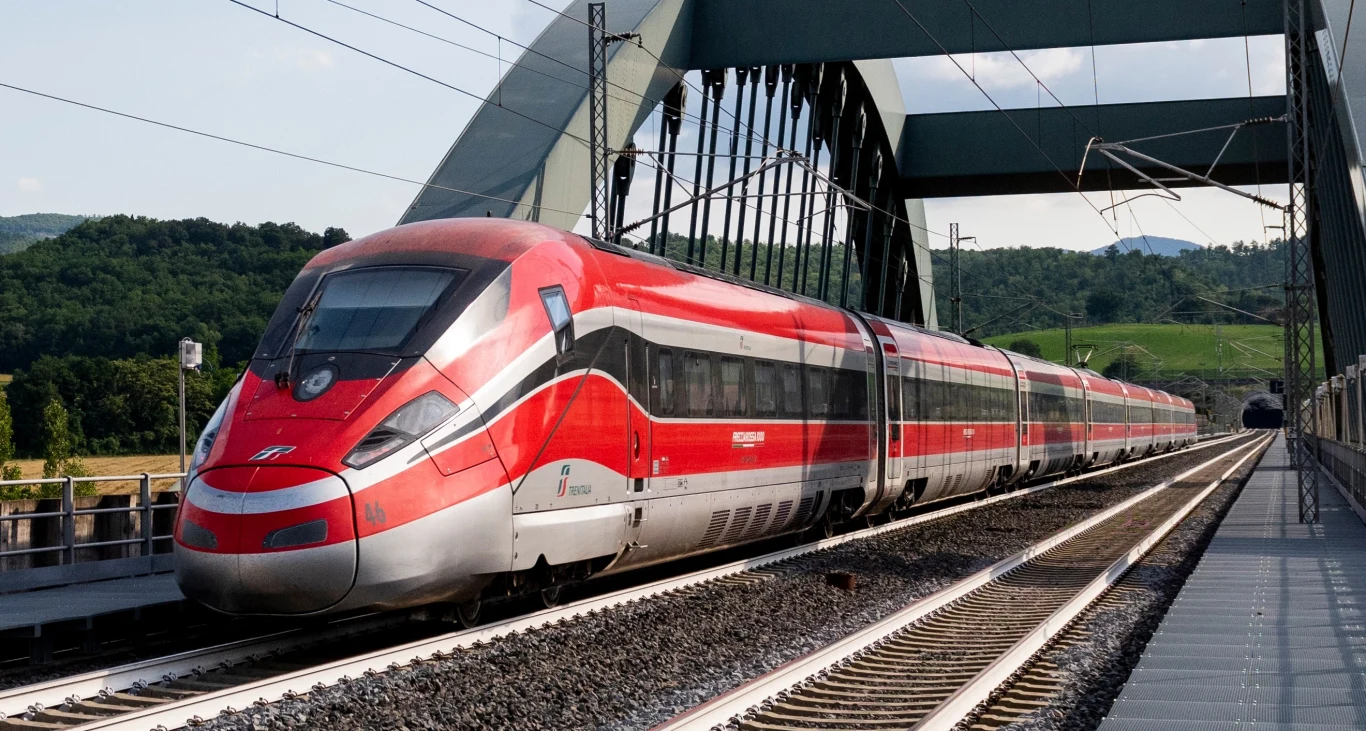 Nowoczesny czerwono-srebrny pociąg dużych prędkości PESA na moście na tle zielonych wzgórz.