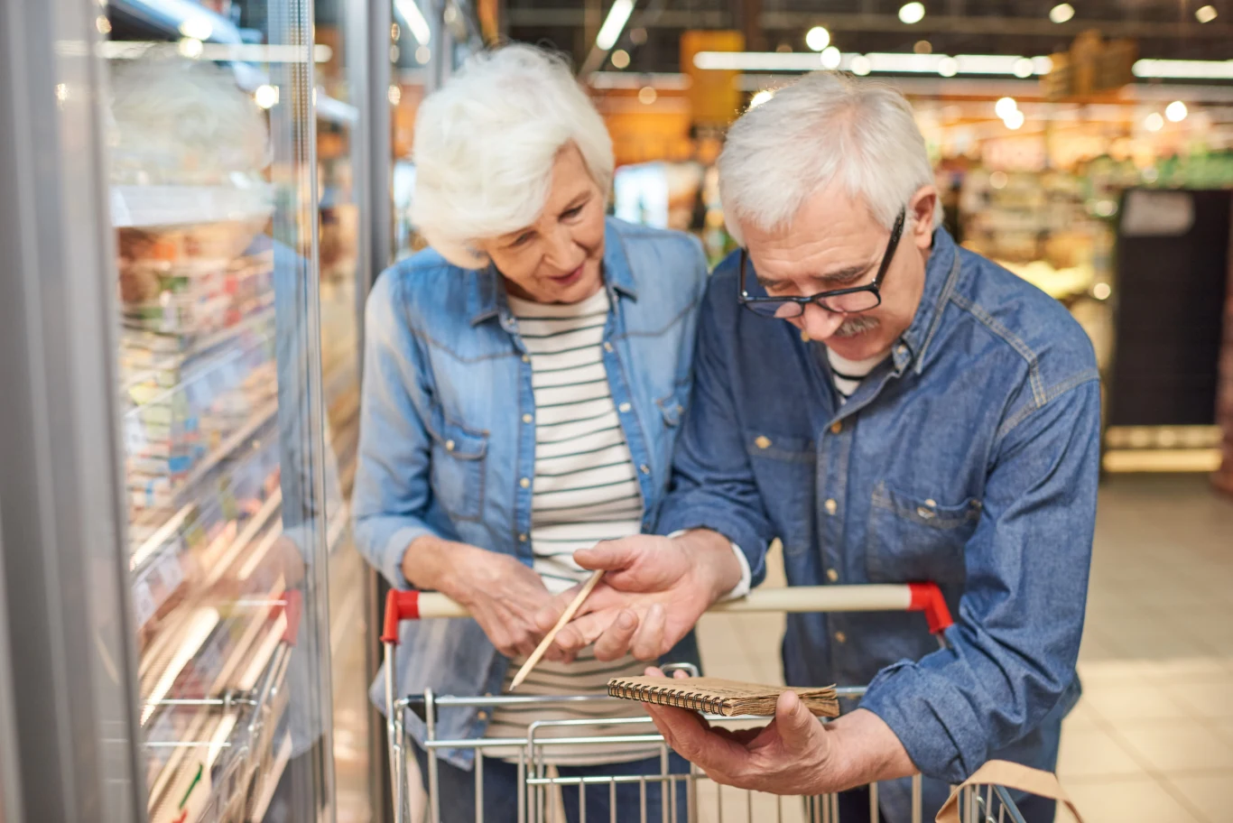 Starsze małżeństwo z wózkiem sklepowym analizujące listę zakupów w supermarkecie.