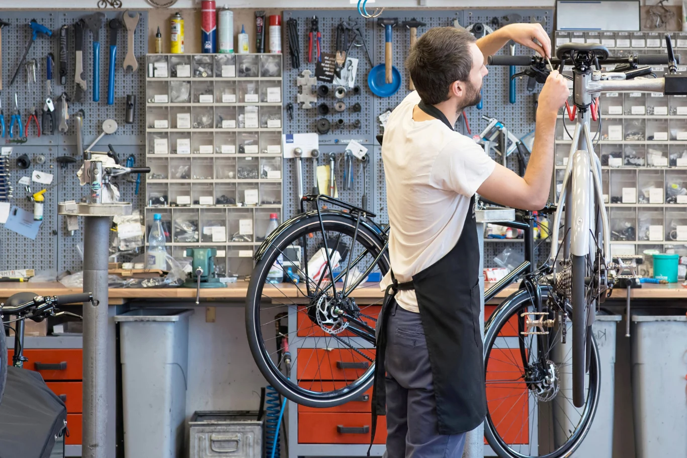 Mechanik w czarnym fartuchu reguluje kierownicę roweru zawieszonego na specjalnym stojaku w dobrze wyposażonym warsztacie rowerowym, otoczony zestawami narzędzi i częściami zamiennymi.
