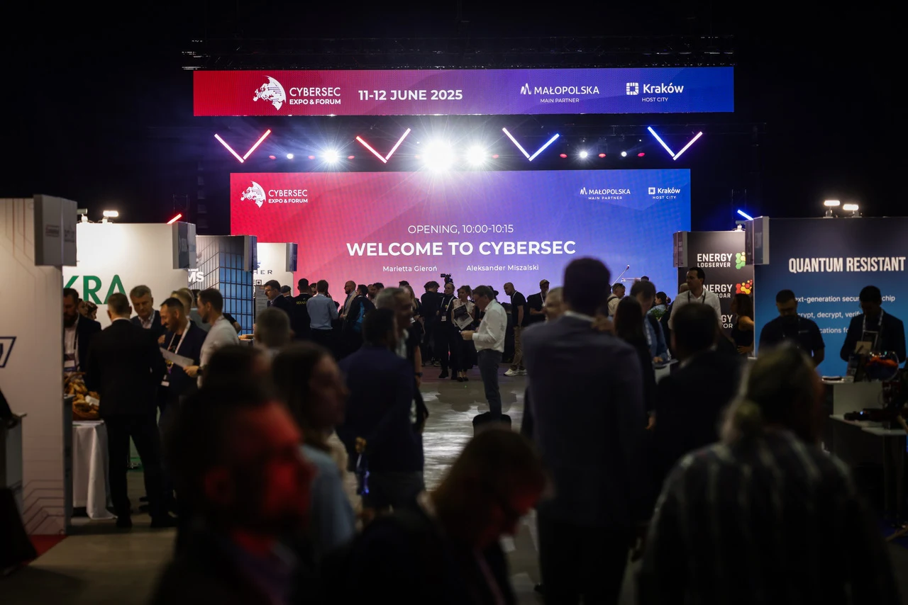 Tłum uczestników konferencji CYBERSEC na tle dużego ekranu z informacją o wydarzeniu i datą, otoczeni stoiskami wystawców związanych z branżą technologii i cyberbezpieczeństwa.
