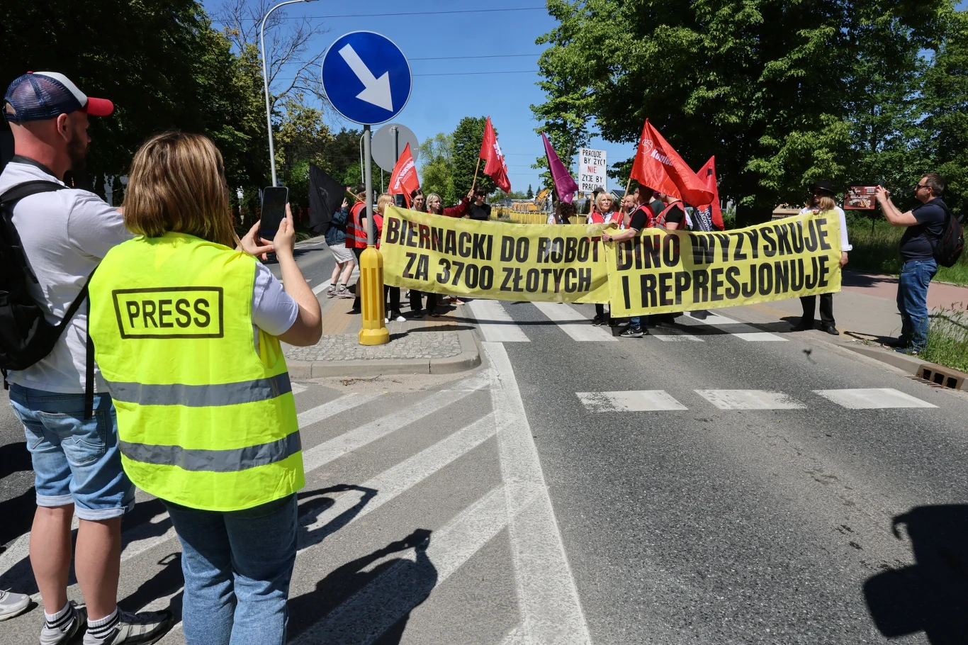 Demonstranci niosą żółte transparenty z hasłami krytykującymi firmę Dino i osobę Biernatckiego, dziennikarka w kamizelce 'PRESS' fotografuje protestujących, w tle widoczne czerwone flagi i zieleń drzew.