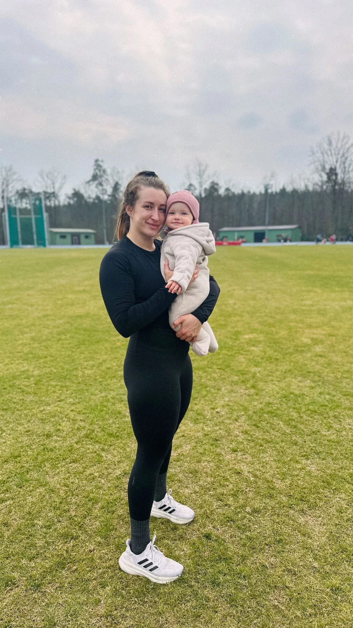 Kobieta w czarnym stroju sportowym trzyma na rękach uśmiechnięte niemowlę ubrane w jasny kombinezon i czapkę, stojąc na zielonym boisku.