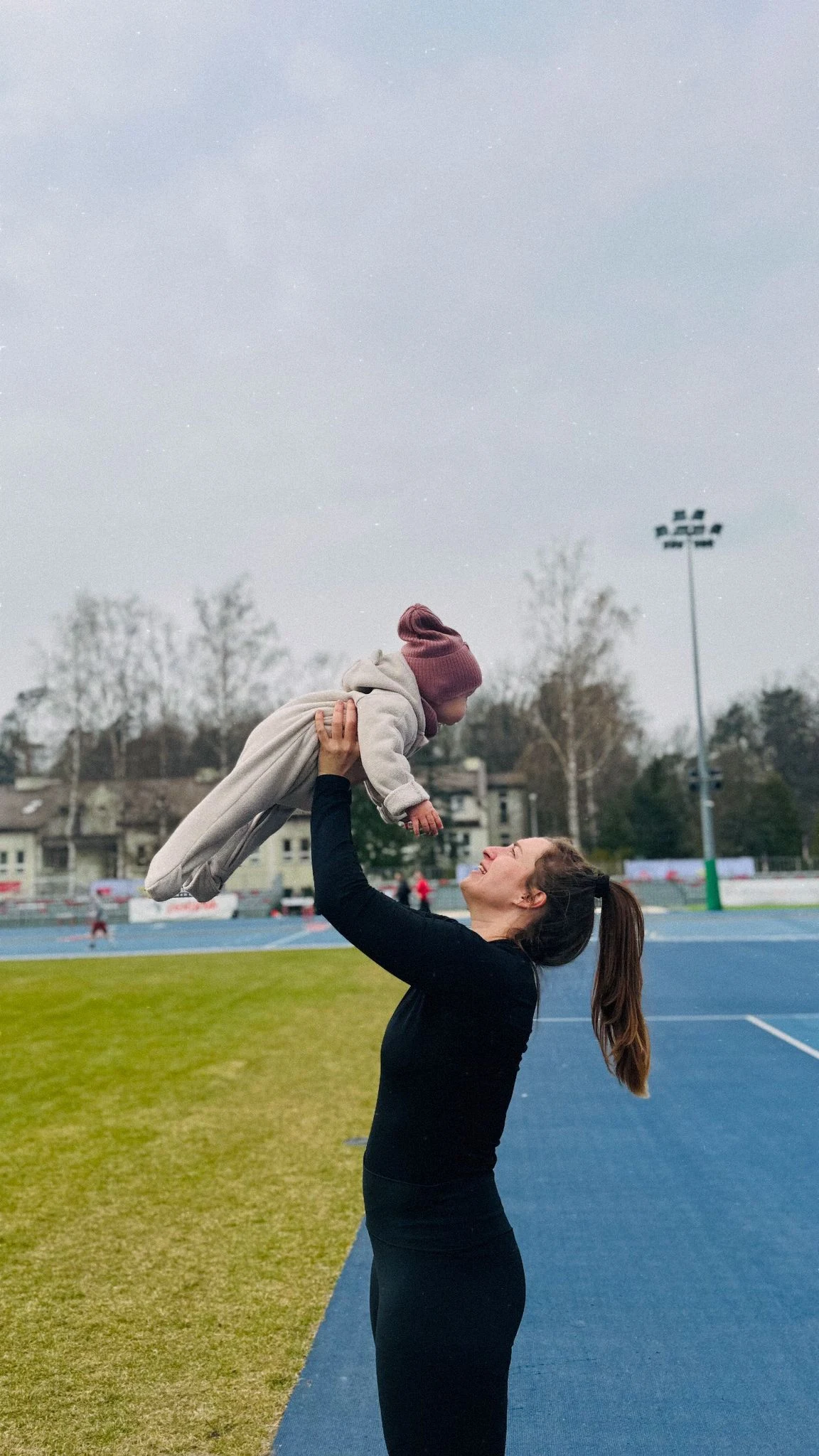 Kobieta w czarnym stroju sportowym unosi uśmiechnięte dziecko ubrane na jasno na tle niebieskiego boiska i zielonej murawy.