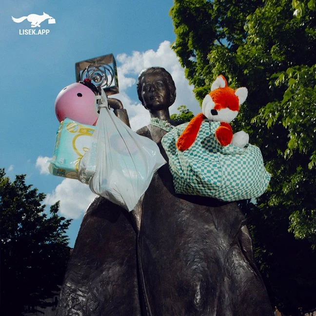 Brązowy pomnik kobiety z zamocowanymi na ramionach torbami wypełnionymi dziecięcymi akcesoriami, w tym pluszowym lisem, pieluchami i różowym kaskiem, na tle zielonych drzew i nieba.