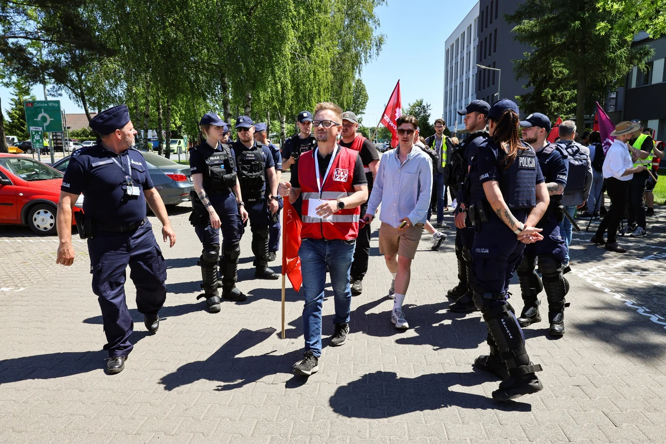 Mężczyzna w czerwonej kamizelce z flagą i megafonem maszeruje w towarzystwie kilku policjantów i innych uczestników demonstracji, wokół widoczne są budynki i drzewa.