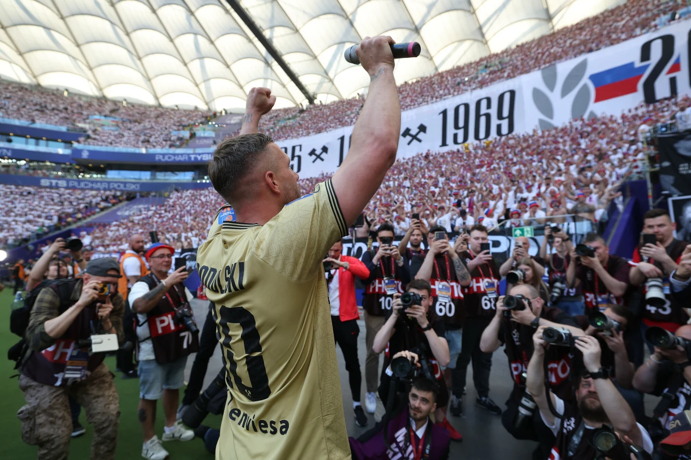 Piłkarz w złotej koszulce z numerem 10 podnosi mikrofon w stronę pełnych trybun, otoczony przez fotografów i kibiców na stadionie.