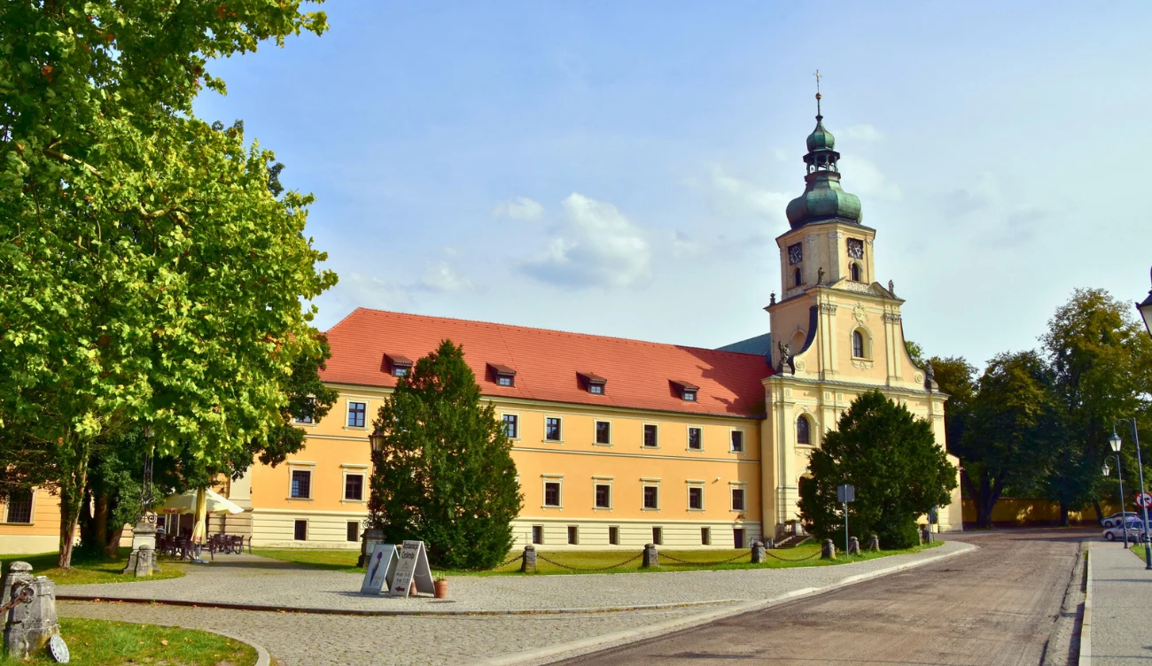 Jasna elewacja zabytkowego budynku z czerwoną dachówką oraz wieżą zwieńczoną zieloną kopułą, otoczenie zieleni i brukowana droga po prawej stronie.