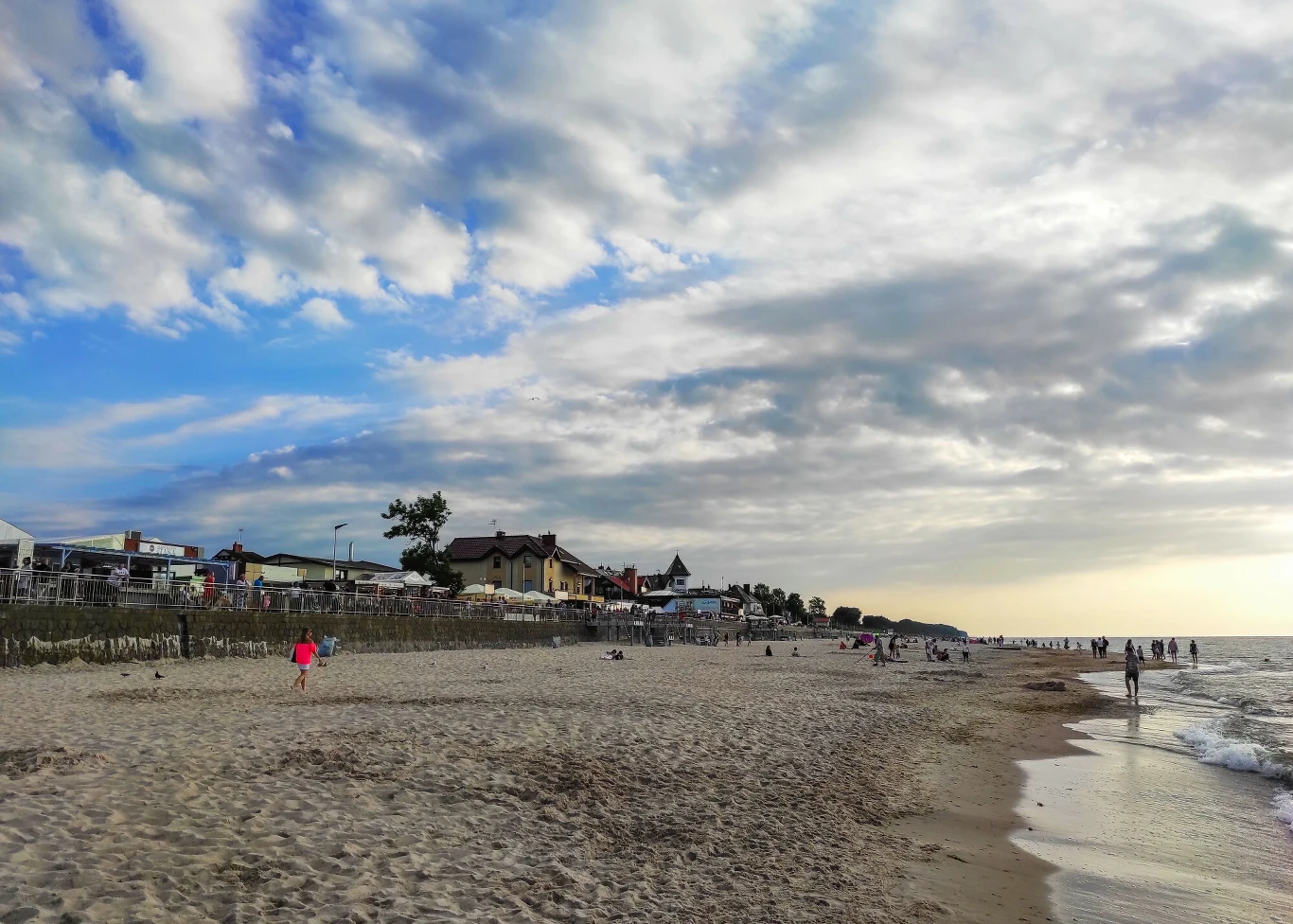 Sarbinowo Piaszczysta plaża z kilkoma spacerującymi osobami, spokojne morze po prawej stronie oraz zabudowania i ogrodzenia przy wydmach w tle pod zachmurzonym niebem.