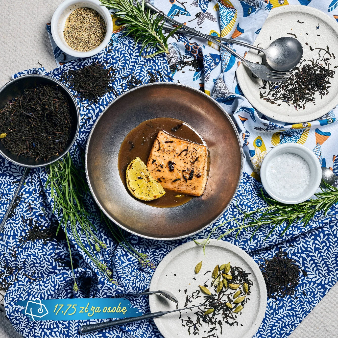 Grillowany kawałek łososia z plasterkiem cytryny w sosie, otoczony świeżymi ziołami oraz przyprawami, ułożony na dekoracyjnym obrusie z niebieskim wzorem i ceramicznymi talerzami.