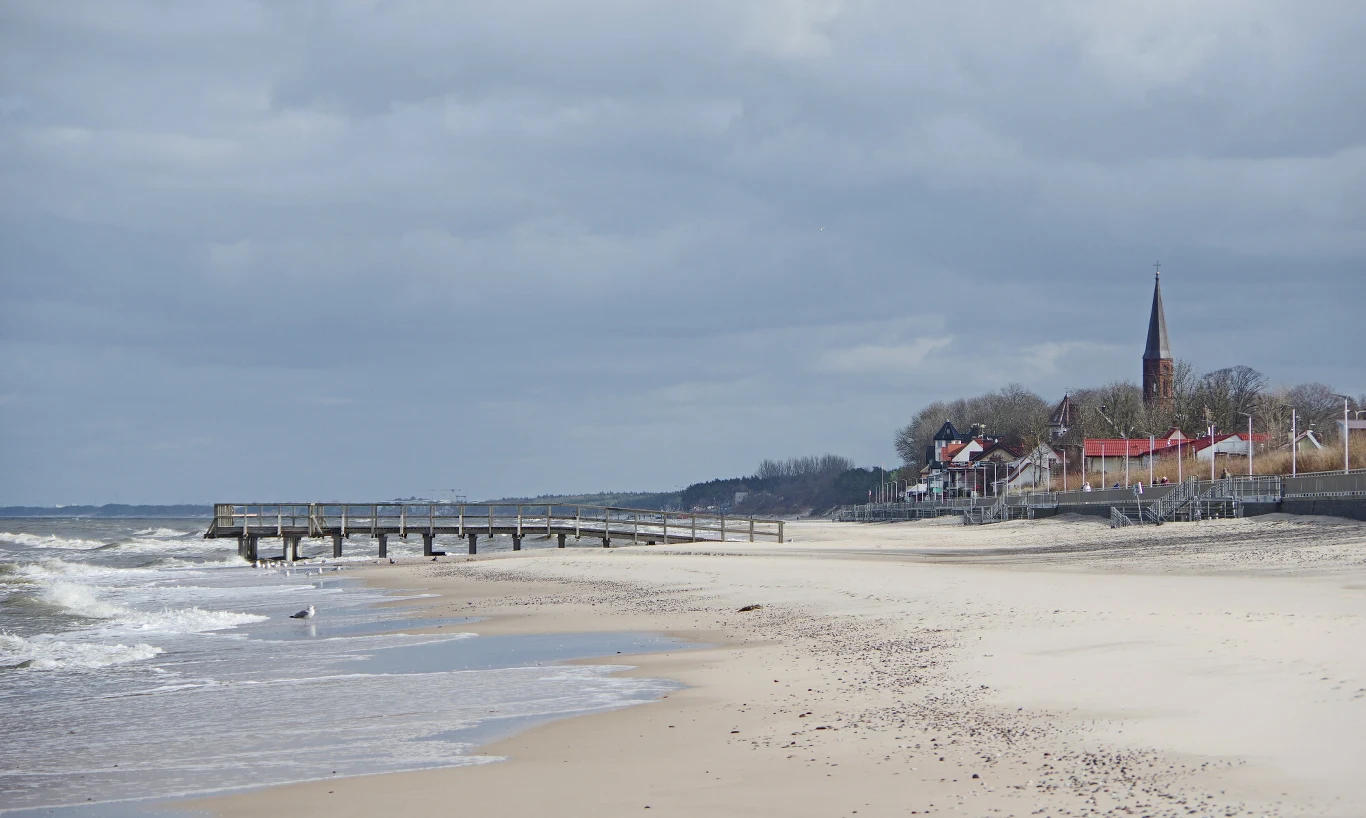 Widok na Sarbinowo od strony morza Piaszczysta plaża z molo, spokojne fale Morza Bałtyckiego, w oddali rząd domów i wysoka wieża kościoła na tle pochmurnego nieba.