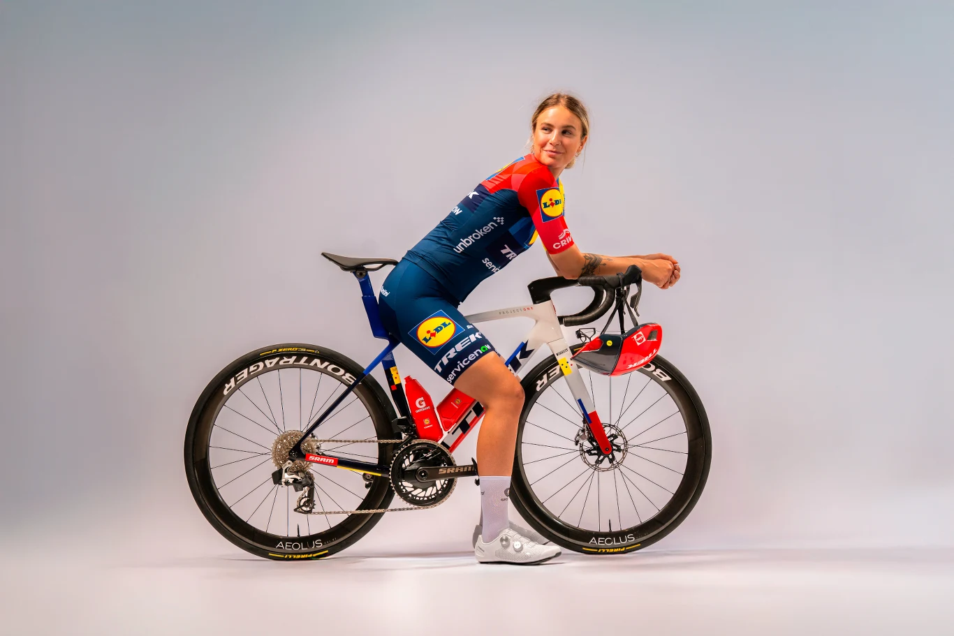 Sprint-rowery.pl Kobieta w stroju kolarskim siedzi na profesjonalnym rowerze szosowym, patrząc w stronę obiektywu; na stroju i rowerze widoczne kolorowe logotypy sponsorskie.