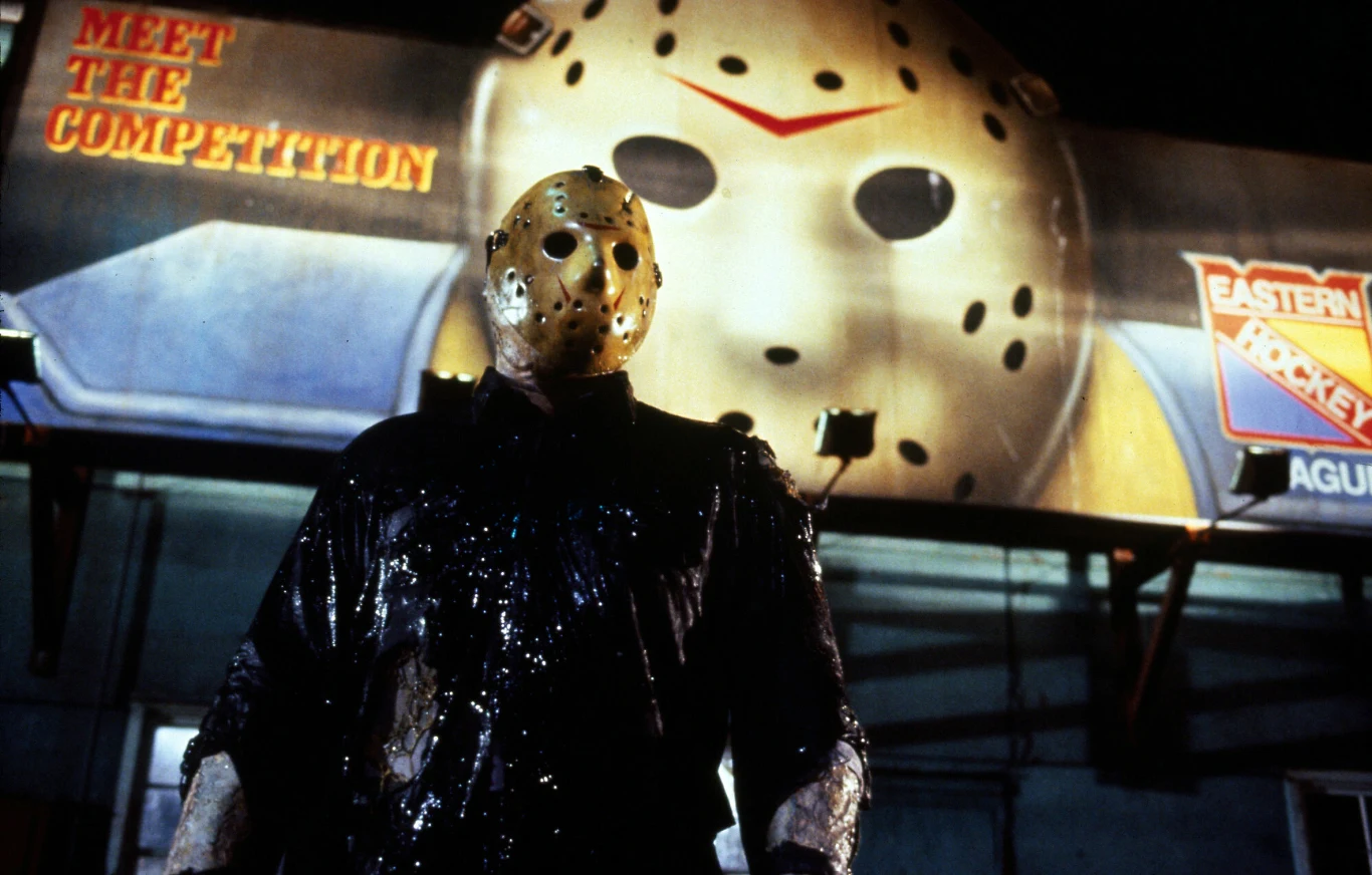Jason Voorhees Jason Voorhees w czarnej odzieży i hokejowej masce stoi przed billboardem z reklamą i maską.