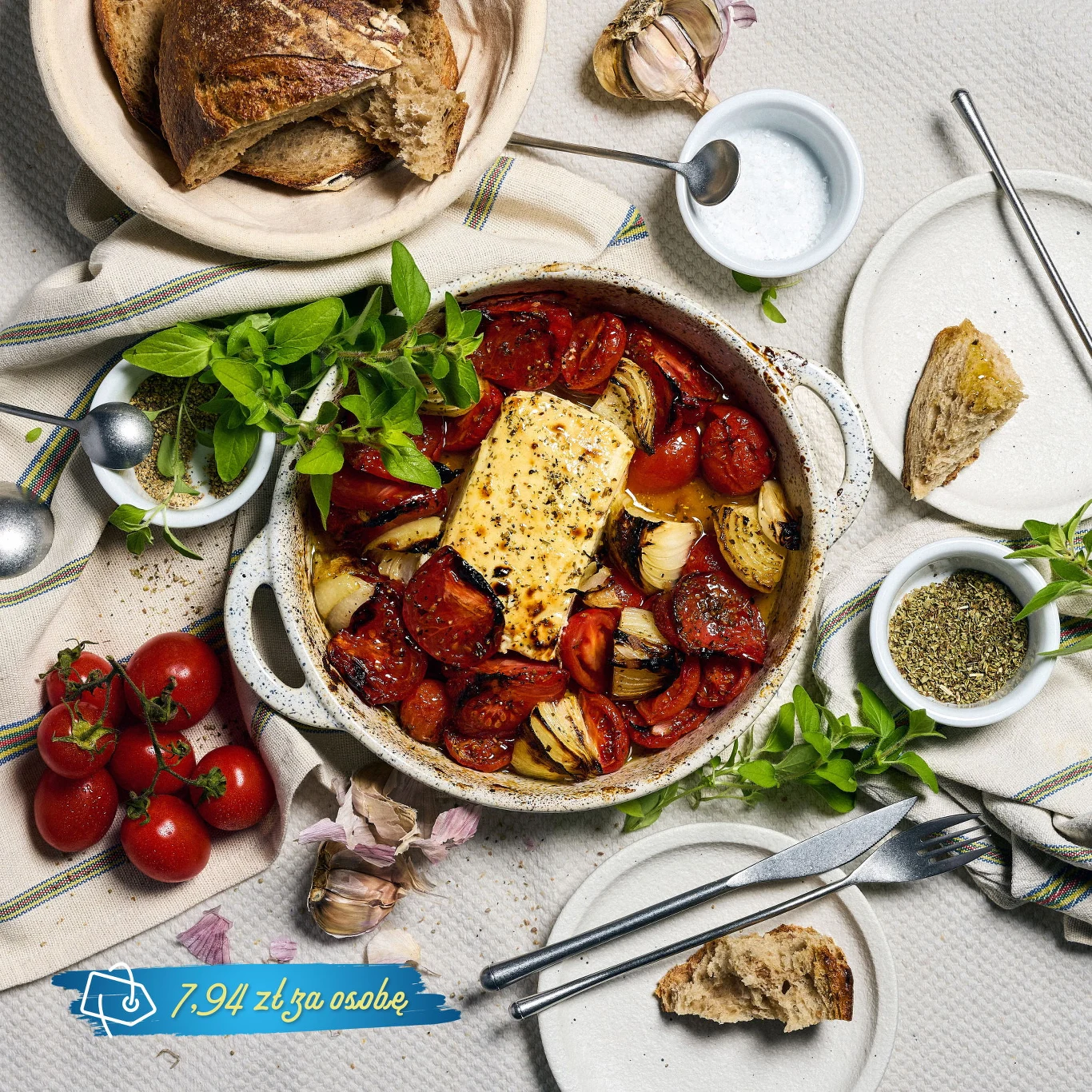 Ułożona w żaroodpornym naczyniu zapiekanka z pieczonymi pomidorami, cebulą i serem feta, udekorowana świeżymi ziołami, otoczona domowym chlebem, czosnkiem i zestawem talerzy oraz sztućców gotowych do podania posiłku.