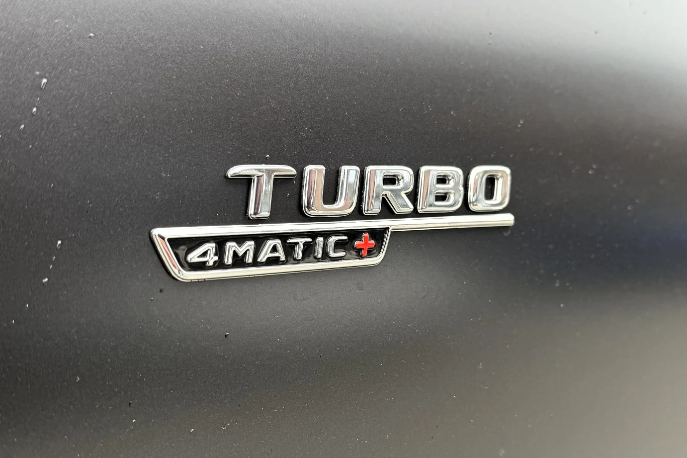 Chromowany emblemat z napisem TURBO 4MATIC+ na czarnym lakierze samochodu.