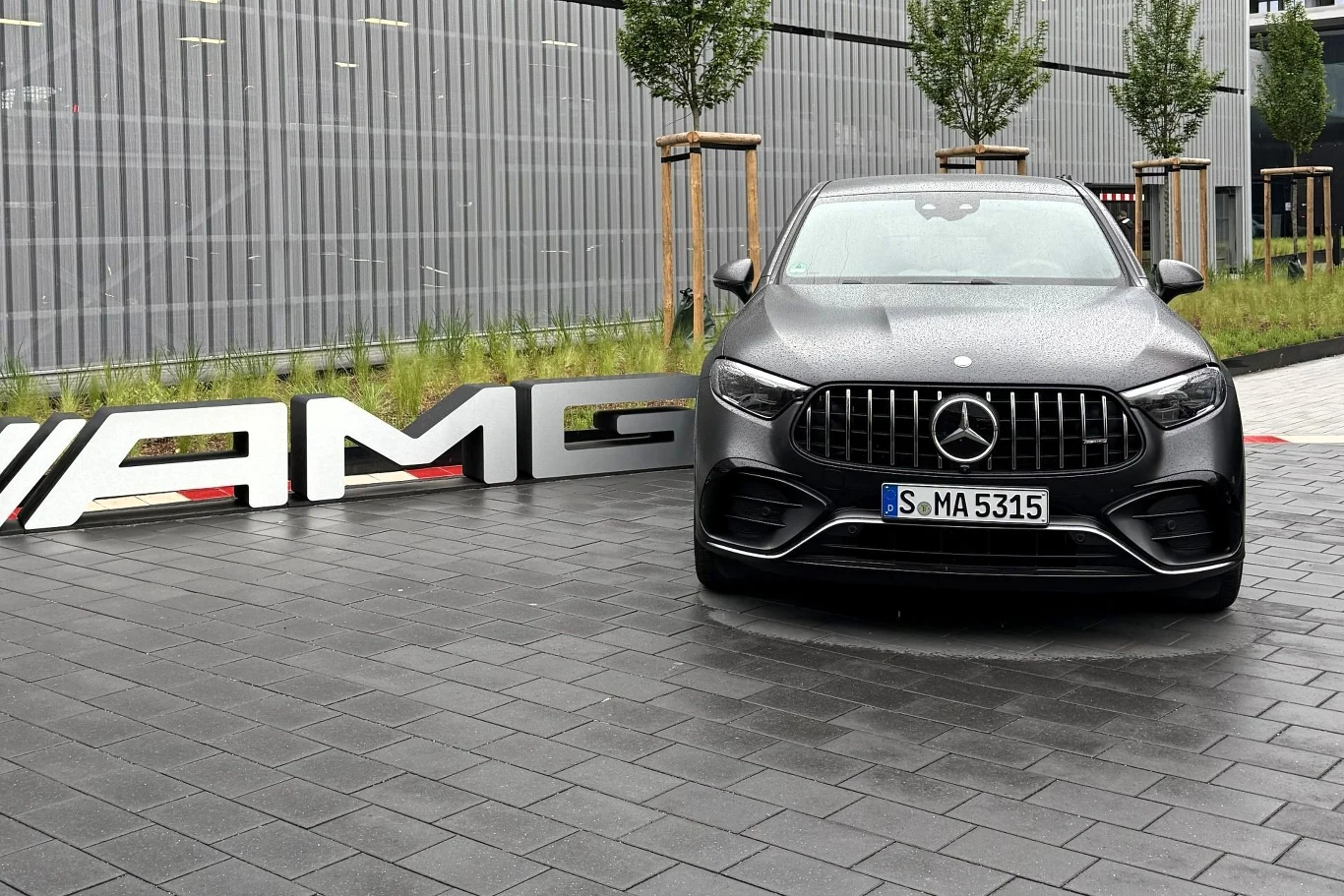 Czarny Mercedes AMG zaparkowany na brukowanej nawierzchni, obok dużego srebrnego napisu AMG, na tle nowoczesnego budynku i kilku młodych drzew.