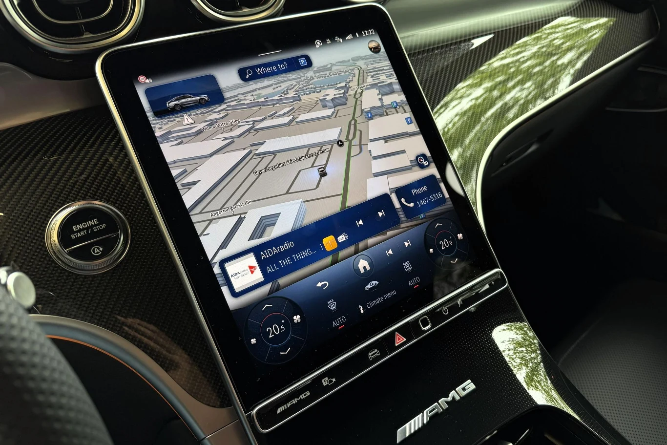 Duży dotykowy ekran systemu multimedialnego z włączoną nawigacją GPS oraz panelem sterowania klimatyzacją i multimediami, otoczony elementami wnętrza z czarnym karbonowym wykończeniem oraz logotypem AMG.