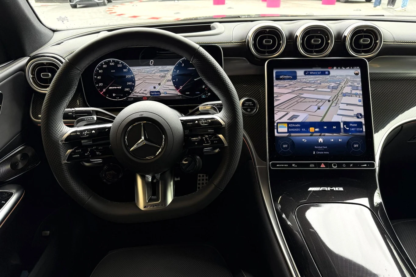 Kokpit samochodu Mercedes-Benz z nowoczesnym wyświetlaczem cyfrowym za kierownicą oraz dużym pionowym ekranem dotykowym z nawigacją po prawej stronie, trzy okrągłe nawiewy powietrza na środku deski rozdzielczej oraz sportowa kierownica z logo