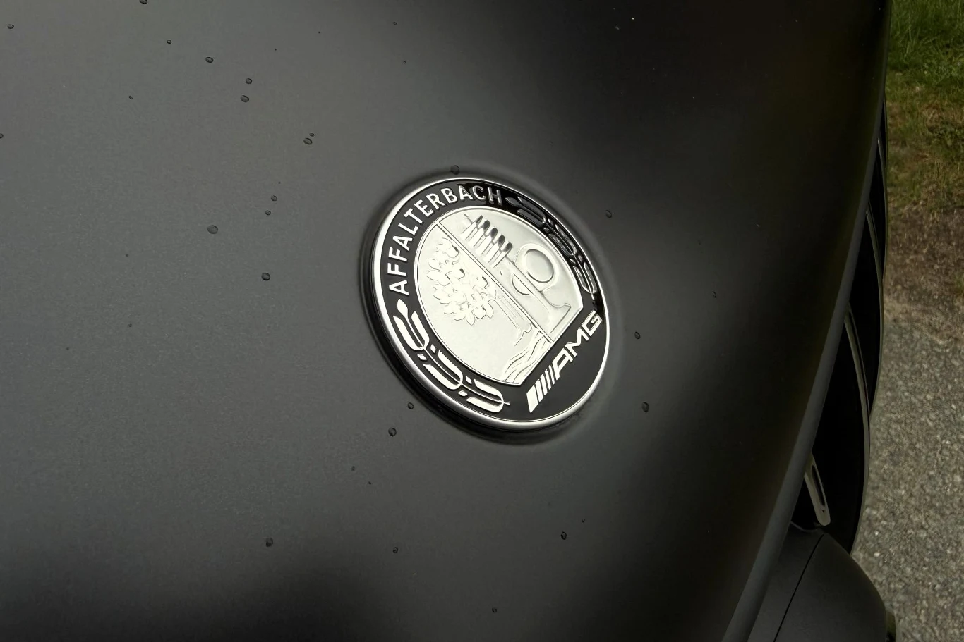 Emblemat AMG z napisem Affalterbach zamontowany na matowoczarnym nadwoziu samochodu, z delikatnymi kroplami wody na powierzchni lakieru.