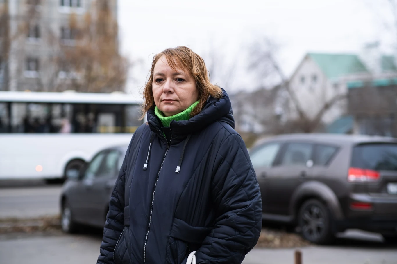 Kobieta w ciemnej, zimowej kurtce stoi na ulicy, w tle zaparkowane samochody i jadący biały autobus.